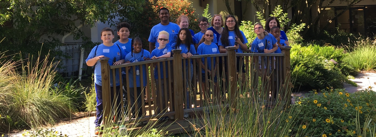 Ten campers and five administrators wearing blue shirts stand on a wooden bridge surrounded by flowers and plants.